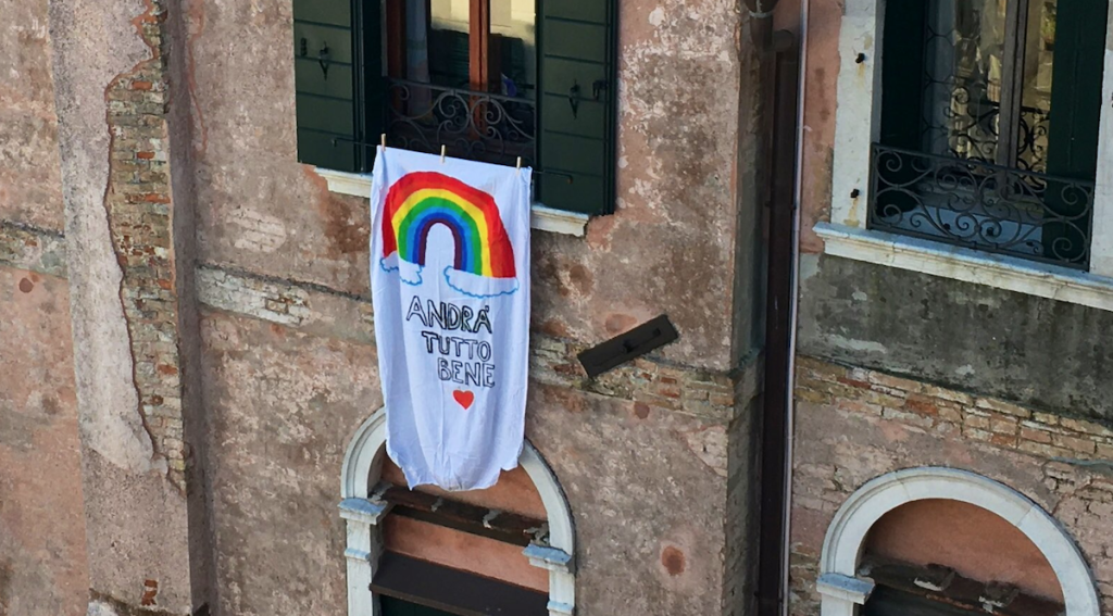 ein selbstgemaltes Plakat hängt aus dem Fenster eines alten Hauses. Darauf ist ein Regenbogen und der Schriftzug „andratuttobene“